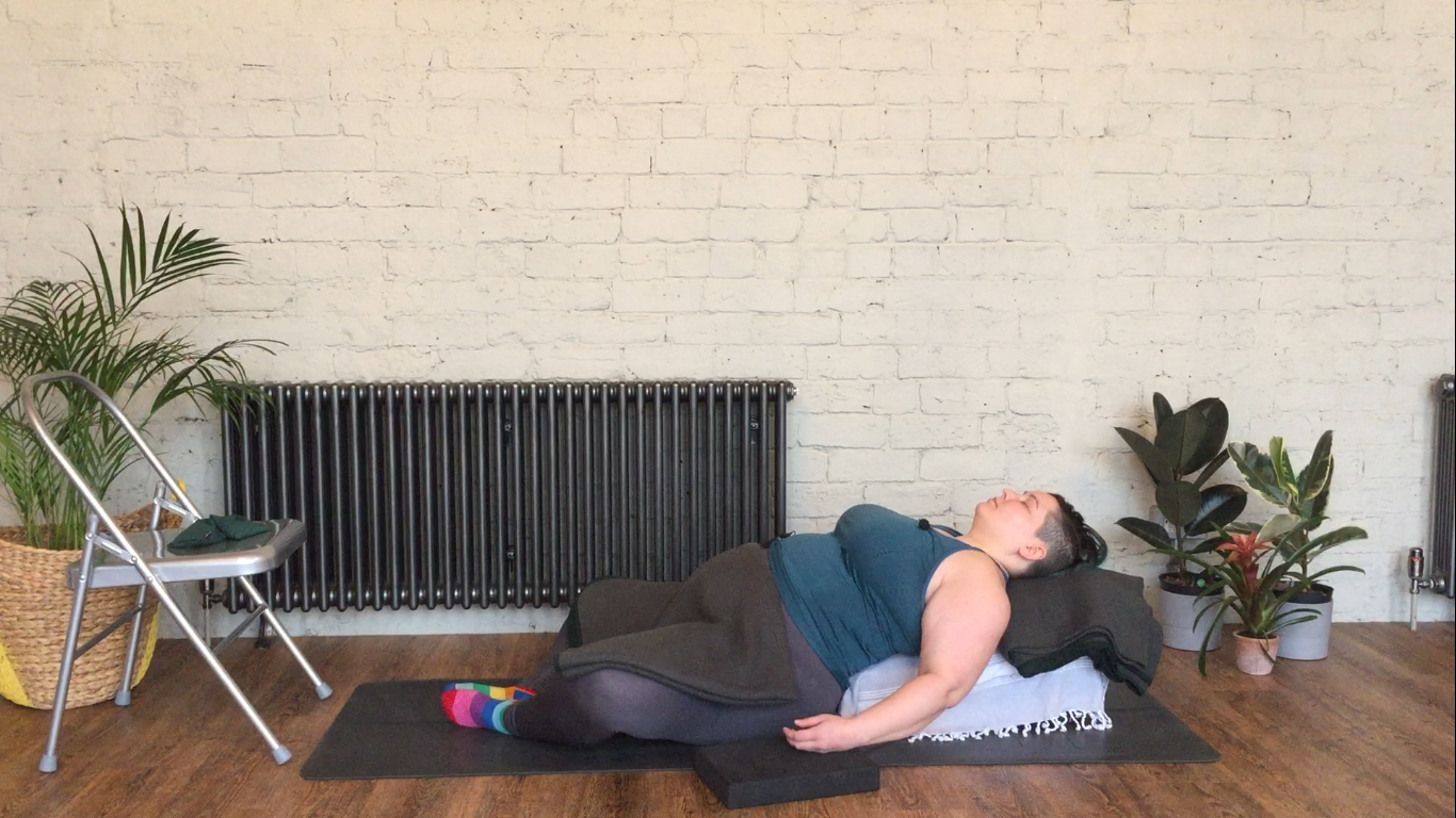 Sarah is laid on a yoga bolster along the spine with feet touching and knees to the side in a reclined butterfly/baddha konasana