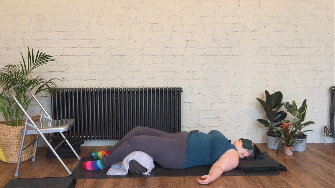 Sarah lays on the ground with a yoga bolster propped under her knees, a blanket under head and an eye pillow over the eyes for savasana