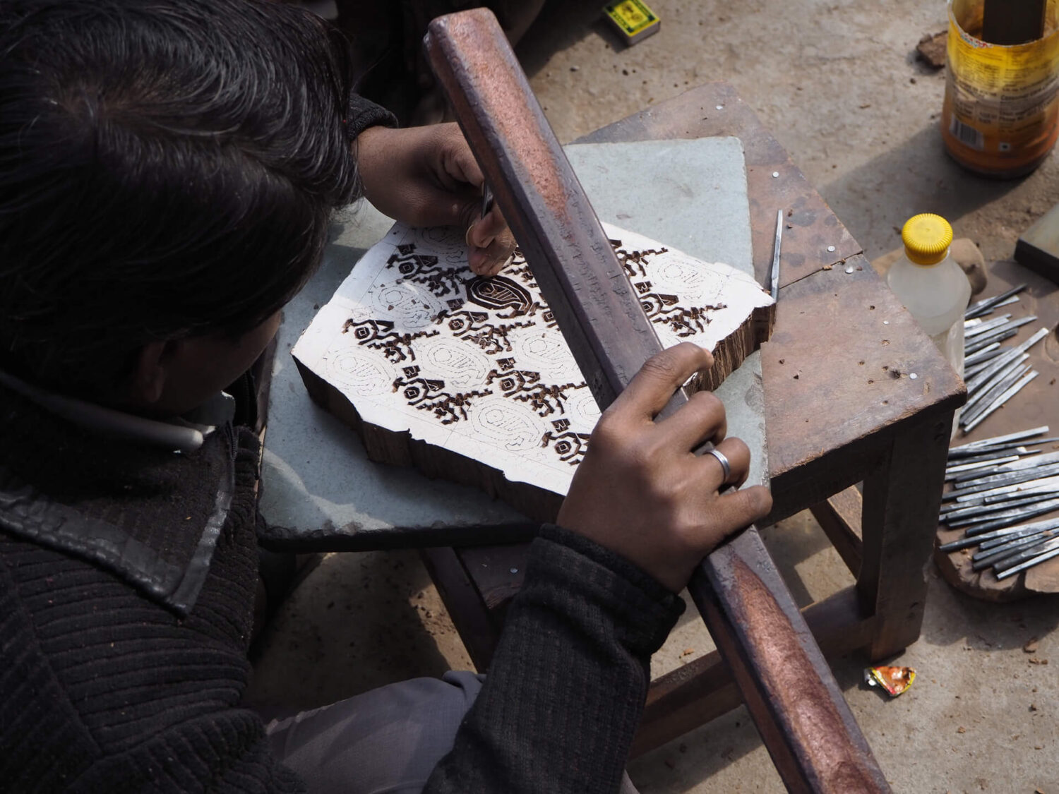 A block carving artisan sits on the ground and hand carves a wooden printing block